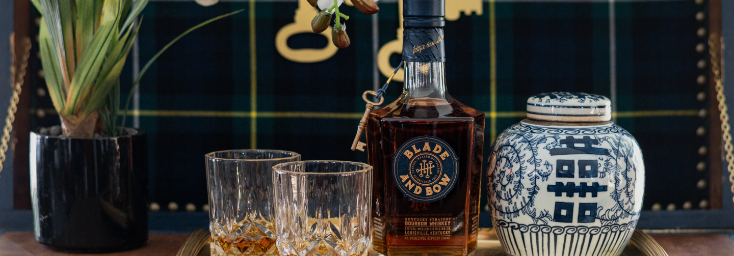 Bottle of Blade and Bow whiskey with an attached key. To the left, two poured highball glasses; decorative vase to the right.