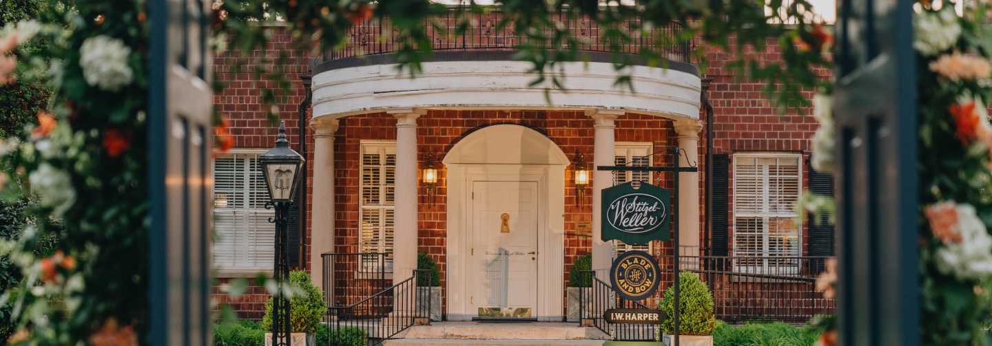 Long shot of the Stitzel-Weller distillery with overhanging branches and gate doors out of focus in the foreground.