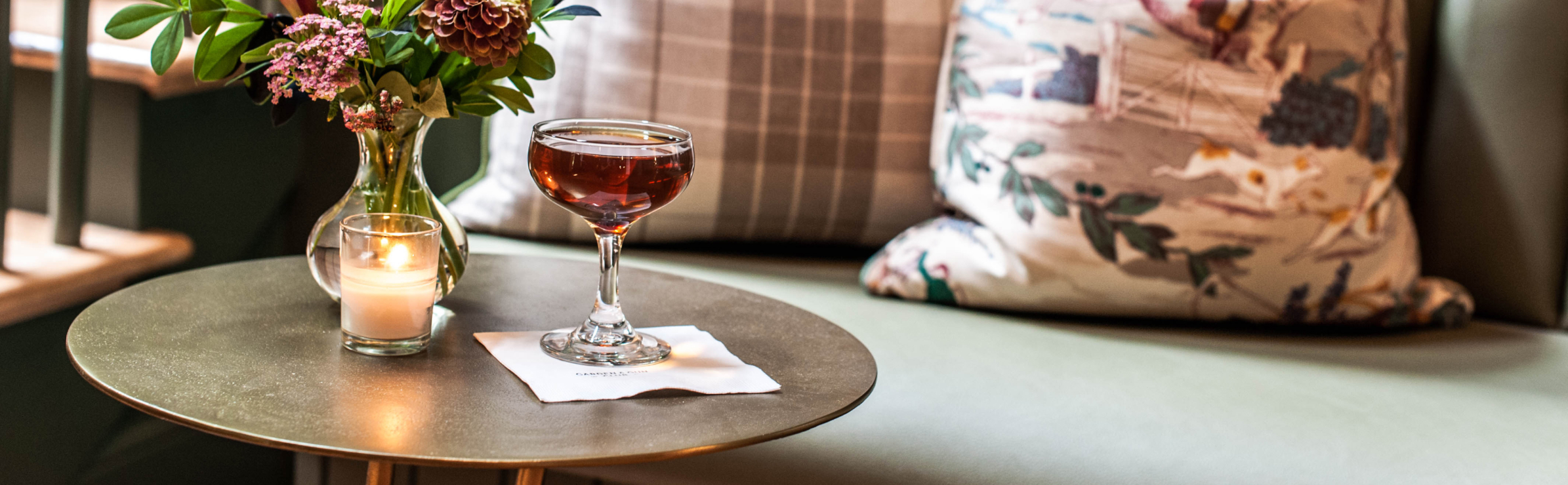 Glass full of Blade and Bow whiskey on an end table with a lit candle and vase of flowers; a couch slightly in the background